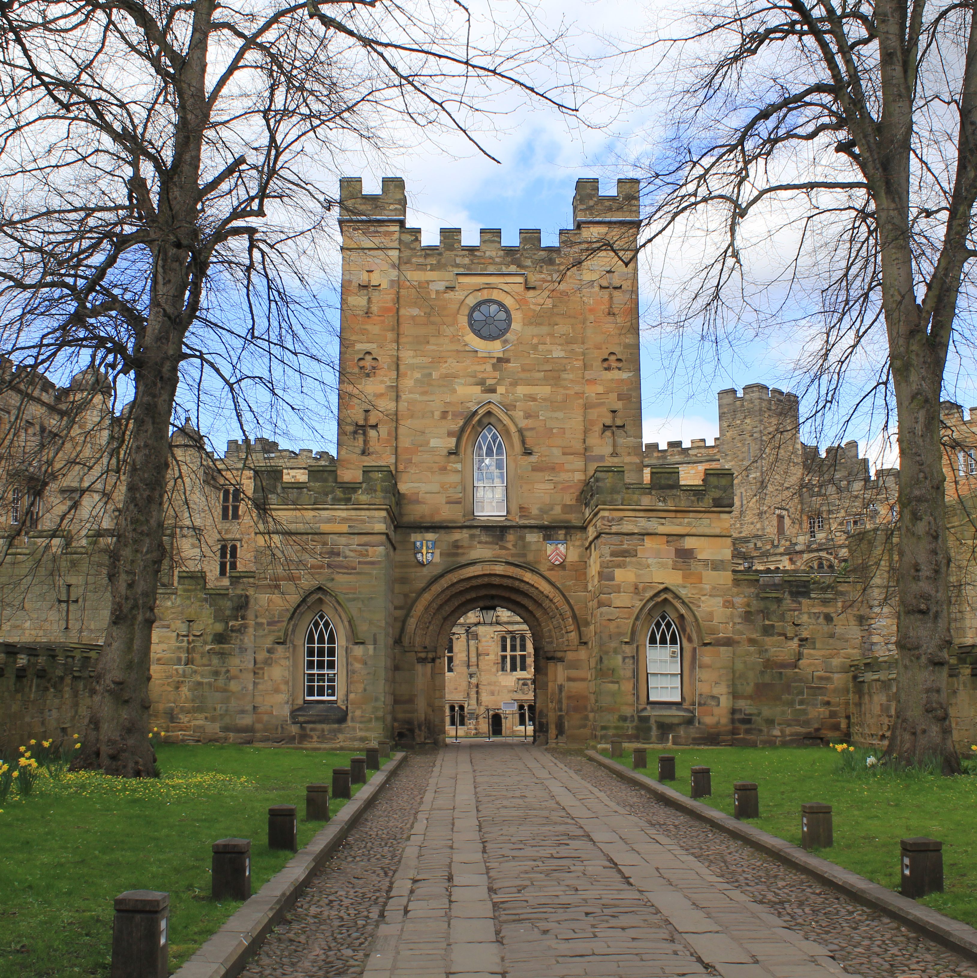 View of Durham Castle, a UNESCO World Heritage Site, showing the historic stone gatehouse with crenellated towers and an arched entrance, framed by leafless trees and a path leading into the castle grounds.