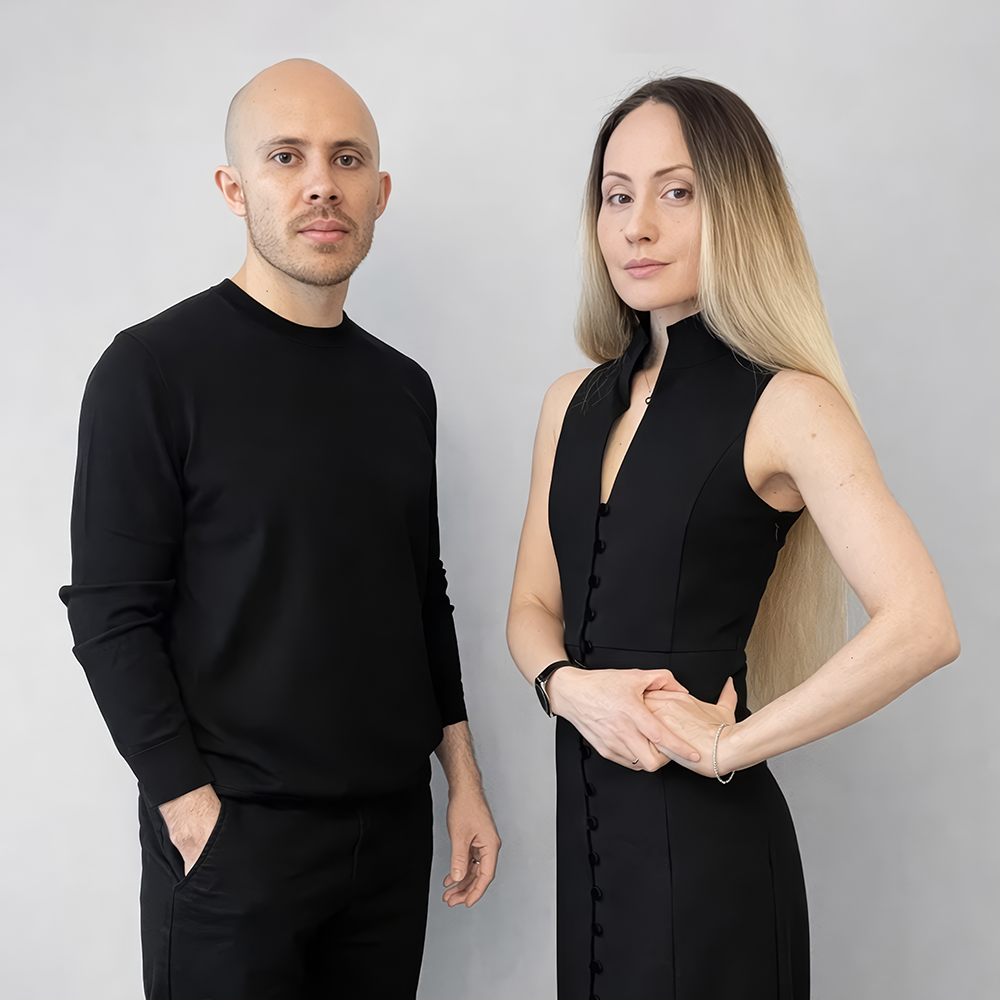 Portrait of A-School alumni Andrew Morrell (left) and Lemara Miftakhova Morrell (right) standing against a neutral background, both dressed in black; Andrew stands with one hand in his pocket, while Lemara faces forward with her hands gently clasped, wearing a sleeveless buttoned dress.
