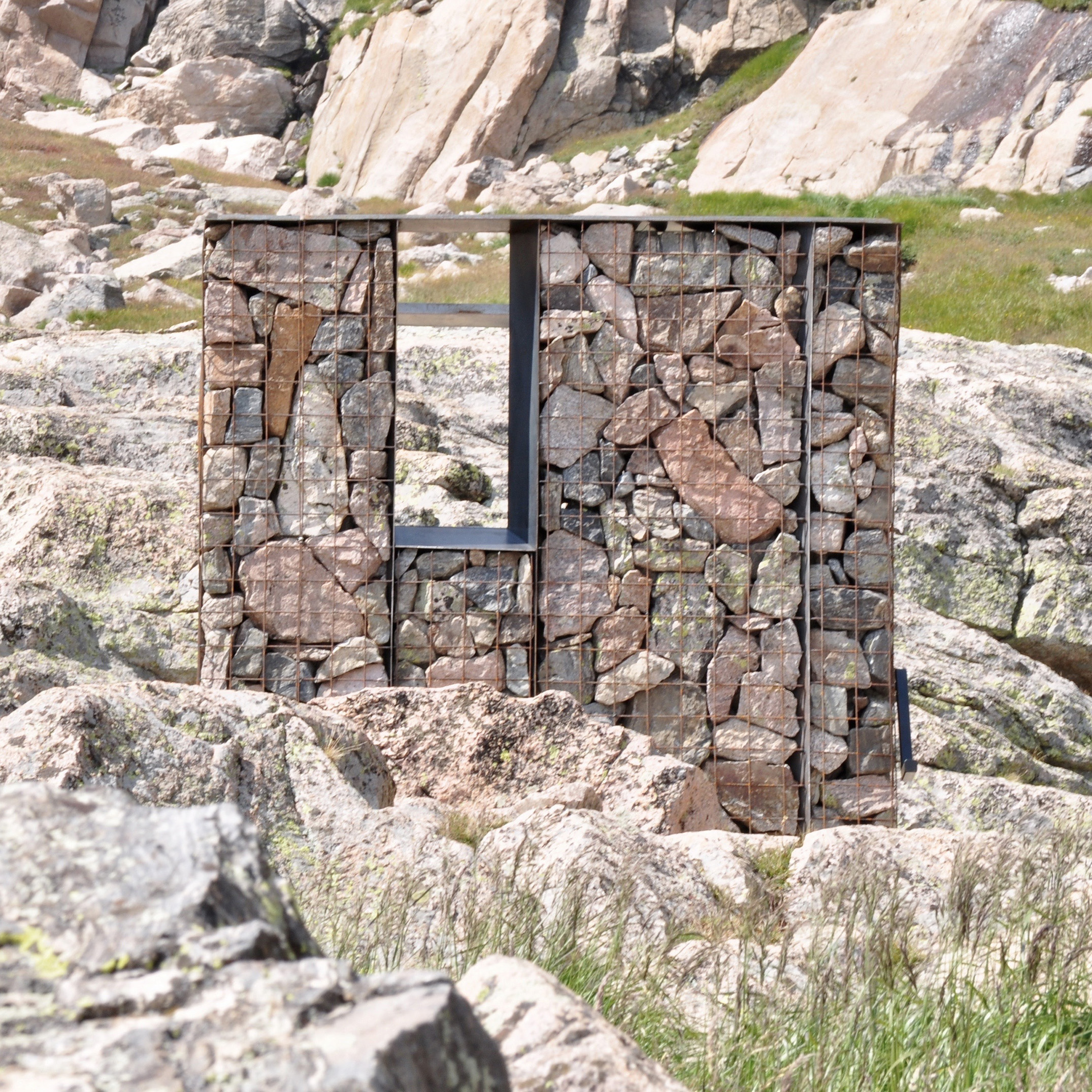 A rectangular gabion structure filled with stones and featuring a square opening stands among sunlit boulders in a rugged alpine environment.