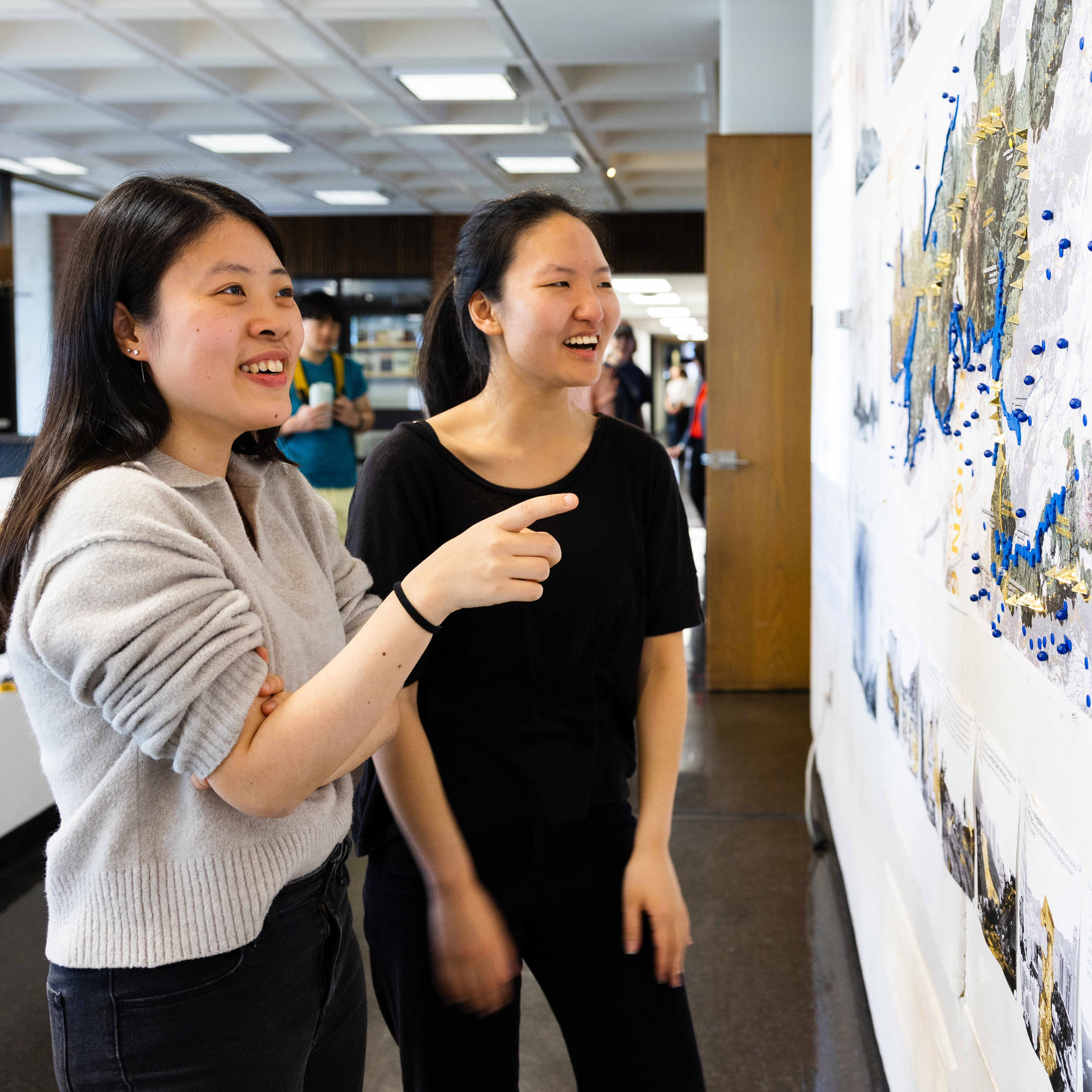Two students laughing and talking to each other while showing each other drawings on the wall