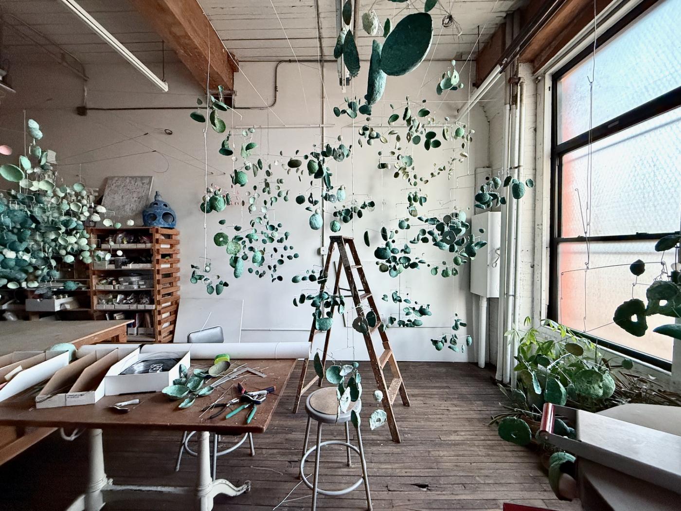 interior of artist's studio filled with hanging sculptural mobiles and ladder in middle of room