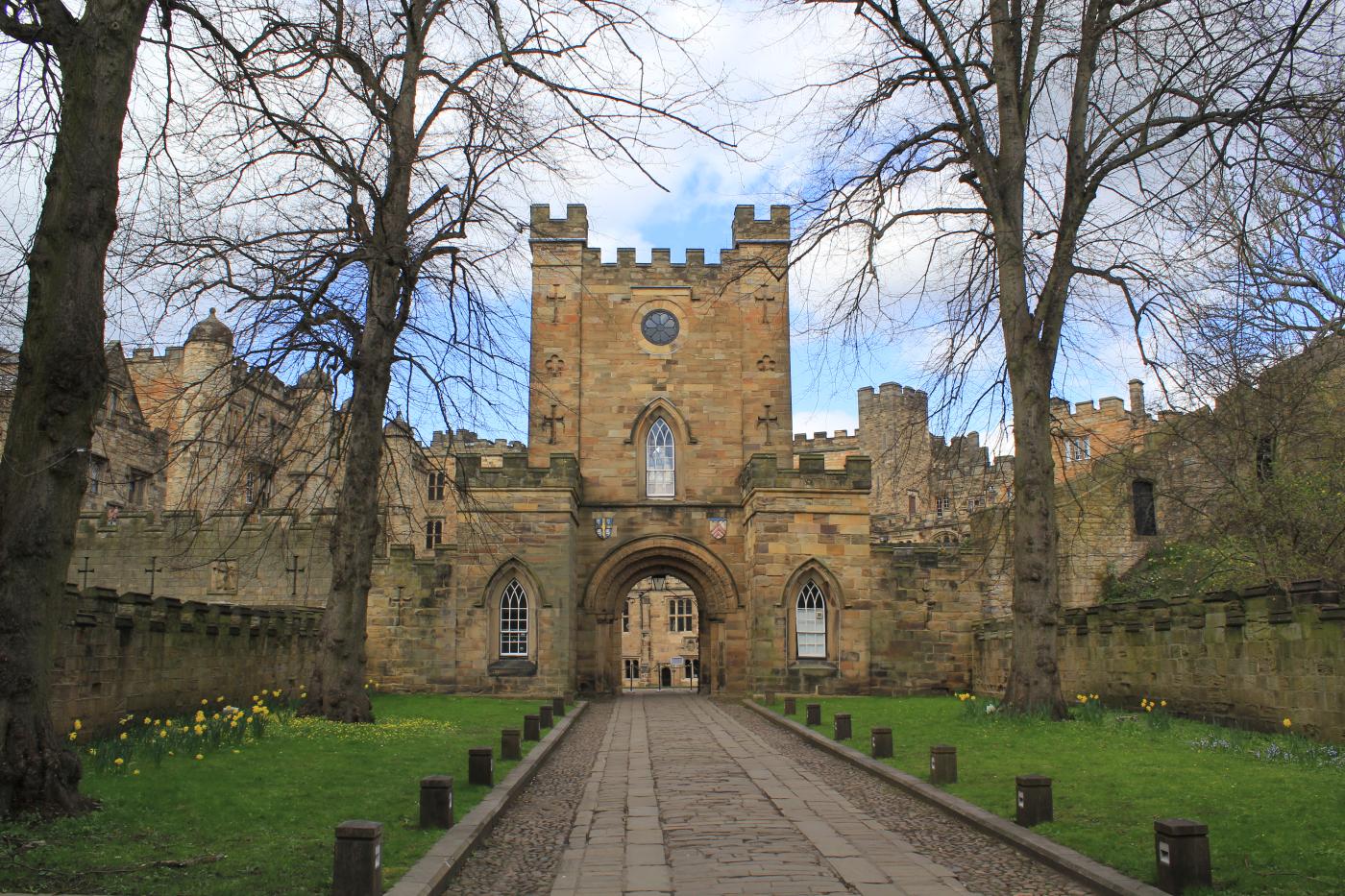View of Durham Castle, a UNESCO World Heritage Site, showing the historic stone gatehouse with crenellated towers and an arched entrance, framed by leafless trees and a path leading into the castle grounds.