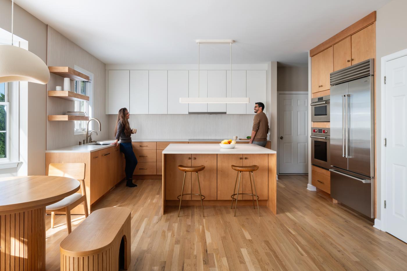 Bright, contemporary kitchen in the Via Florence Renovation featuring warm wood cabinetry, white upper cabinets, and a central island with stools; two people stand at opposite sides of the space, with natural light streaming in from a window and open shelving along the wall.
