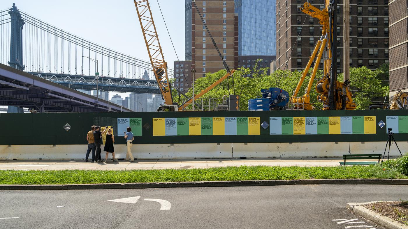 Four people from Citygroup hang colorful posters on construction fence surrounding an urban development site in New York City.