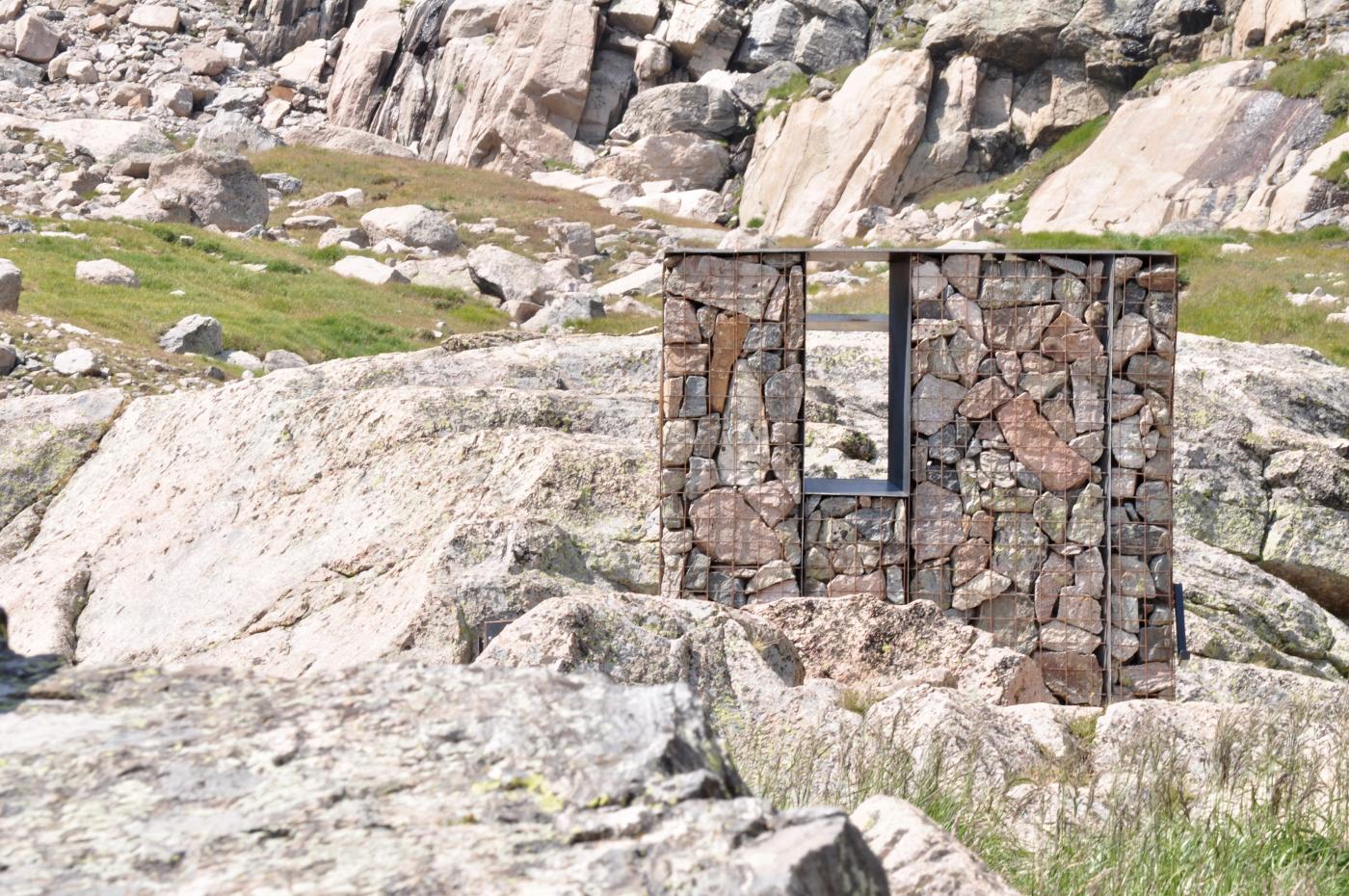 A rectangular gabion structure filled with stones and featuring a square opening stands among sunlit boulders in a rugged alpine environment.