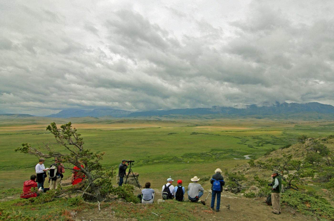 Scenic vista of an expansive valley with mountains in the distance and a dramatic cloudy sky. A group of people sit and stand in the foreground.