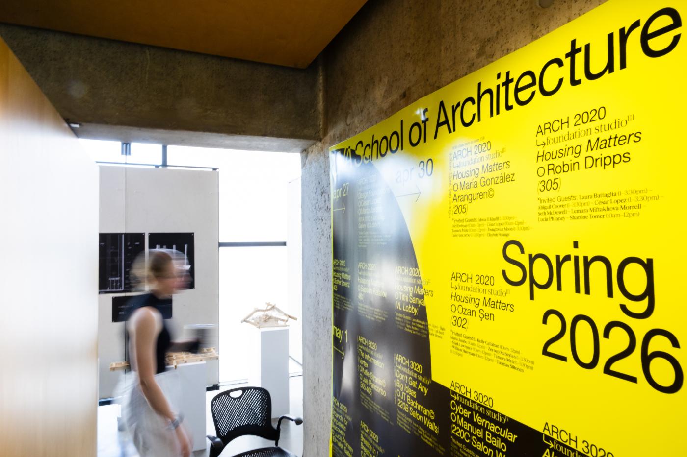 Image of poster of Spring 2026 Final Reviews with person standing in front of student work on display